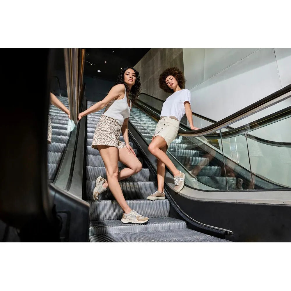 Two women on an escalator wearing beige and white casual shoes, with casual summer outfits, in a modern indoor setting.