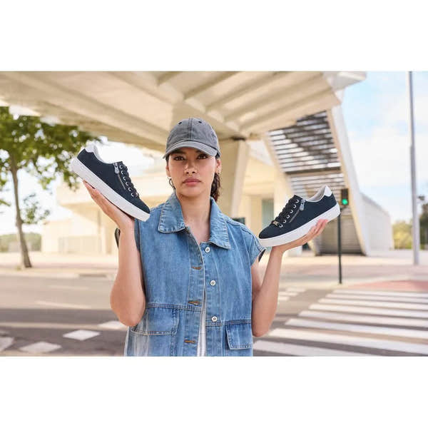 Woman holding black suede sneakers with white soles, lace and zip details, in front of modern architecture.