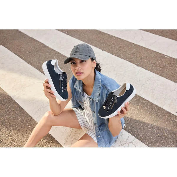 Woman sitting on street crosswalk holding navy blue suede lace-up shoes with side zippers and white soles.