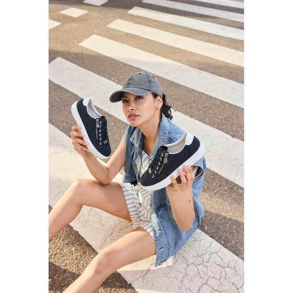 Woman sitting on crosswalk holding black and white suede sneakers with zip and lace details, wearing a gray cap, denim vest, and striped shorts.