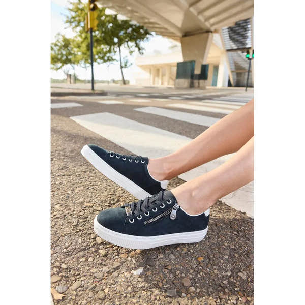 Black suede sneakers with white soles, lace-up front, and side zipper detail, worn outdoors on asphalt.