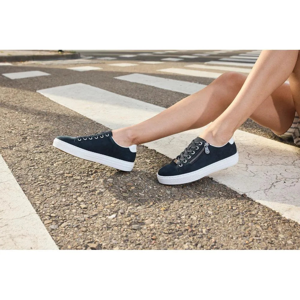 Black suede lace-up shoes with white soles, zip detail on the side, on gravel near a pedestrian crossing.