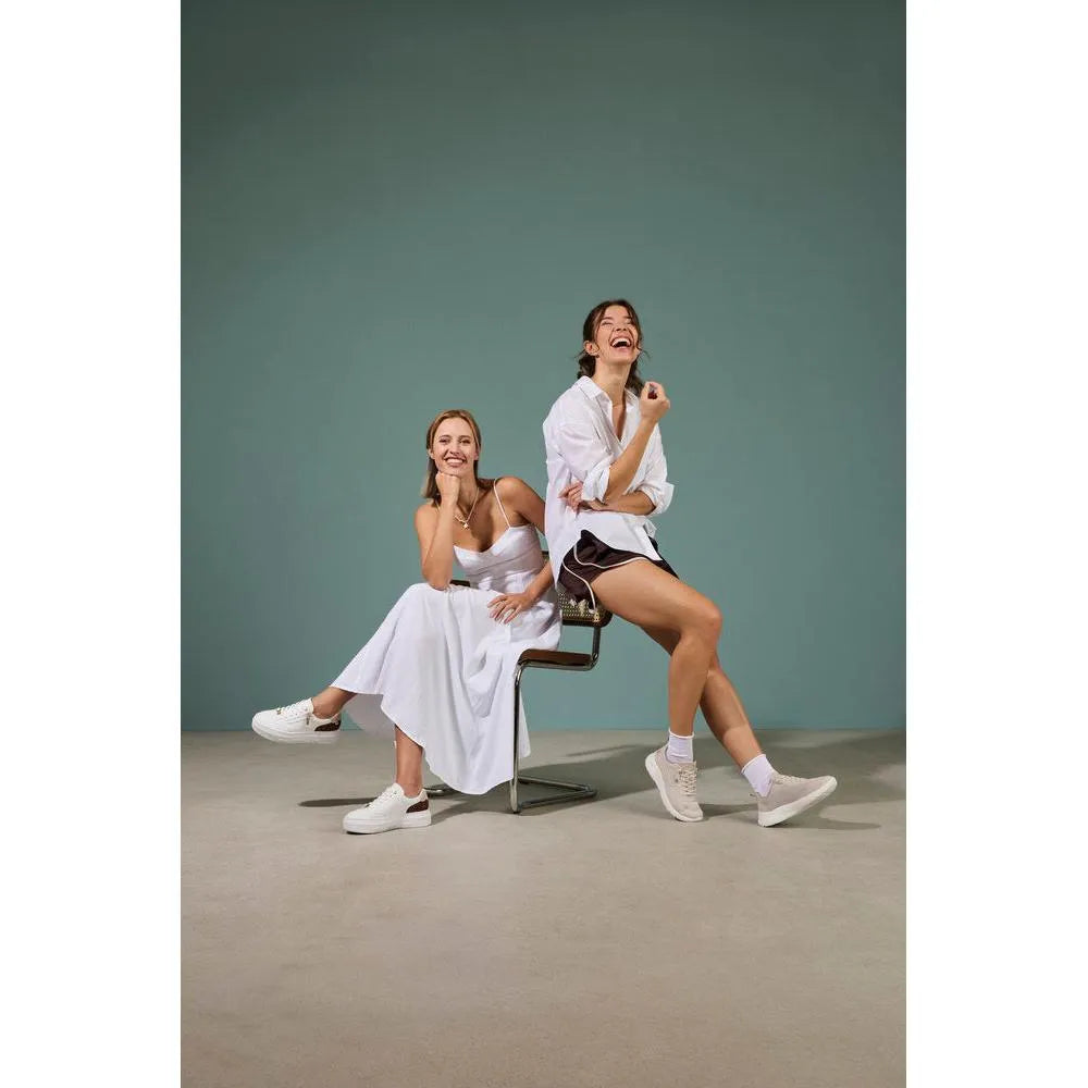 Two women sitting on a modern chair, wearing white and black casual outfits with white trainers; one in leather leopard print trainers.