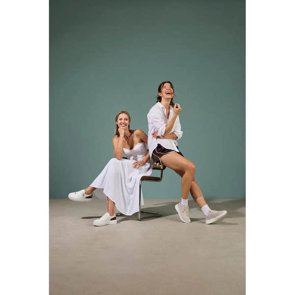 Two women sitting on a modern chair, wearing white and black casual outfits with white trainers; one in leather leopard print trainers.