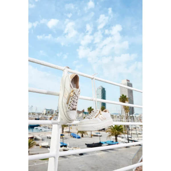 White leather trainers with gold zippers, lace-up detail, and a chunky sole, hanging on a white railing near a marina.