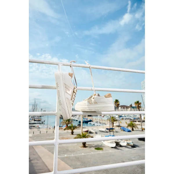 Off-white leather trainers with laces and zippers hanging on a white railing, seaside marina background.