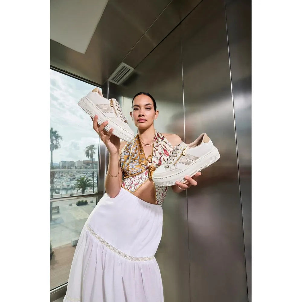 Woman holding off-white leather trainers with zip and lace details, posing indoors with a city view background.
