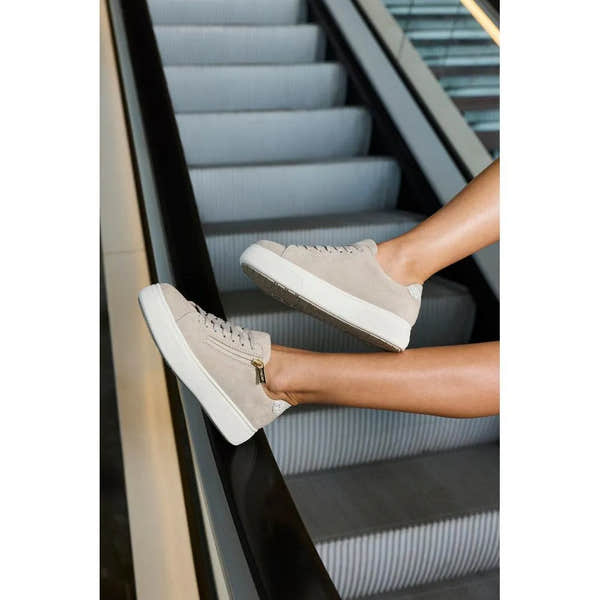 Off-white suede zip and lace women's sneakers on an escalator step, with a thick white sole and subtle textured detail.
