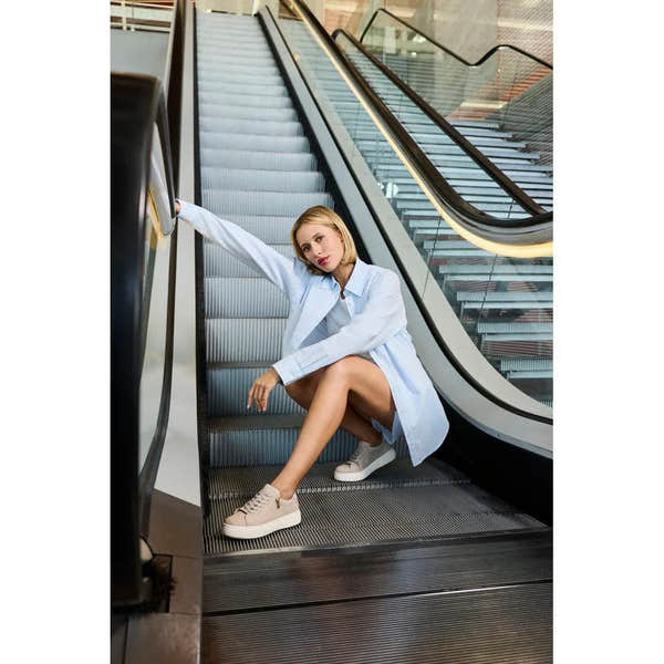 Woman sitting on an escalator wearing off-white suede sneakers with zips and laces, dressed in a light blue shirt.