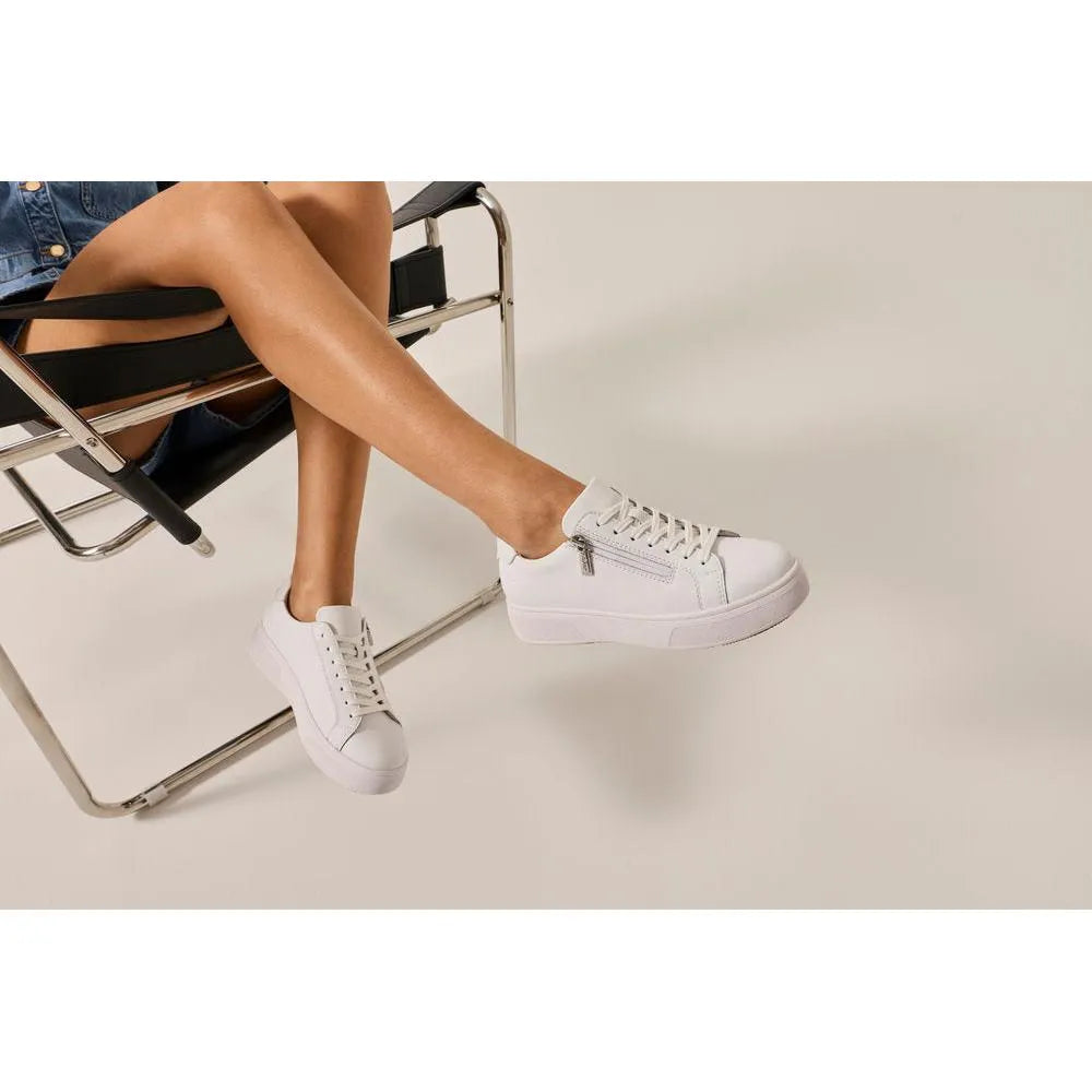 White leather lace-up shoes with side zip detail, worn by a person sitting on a modern black and chrome chair.