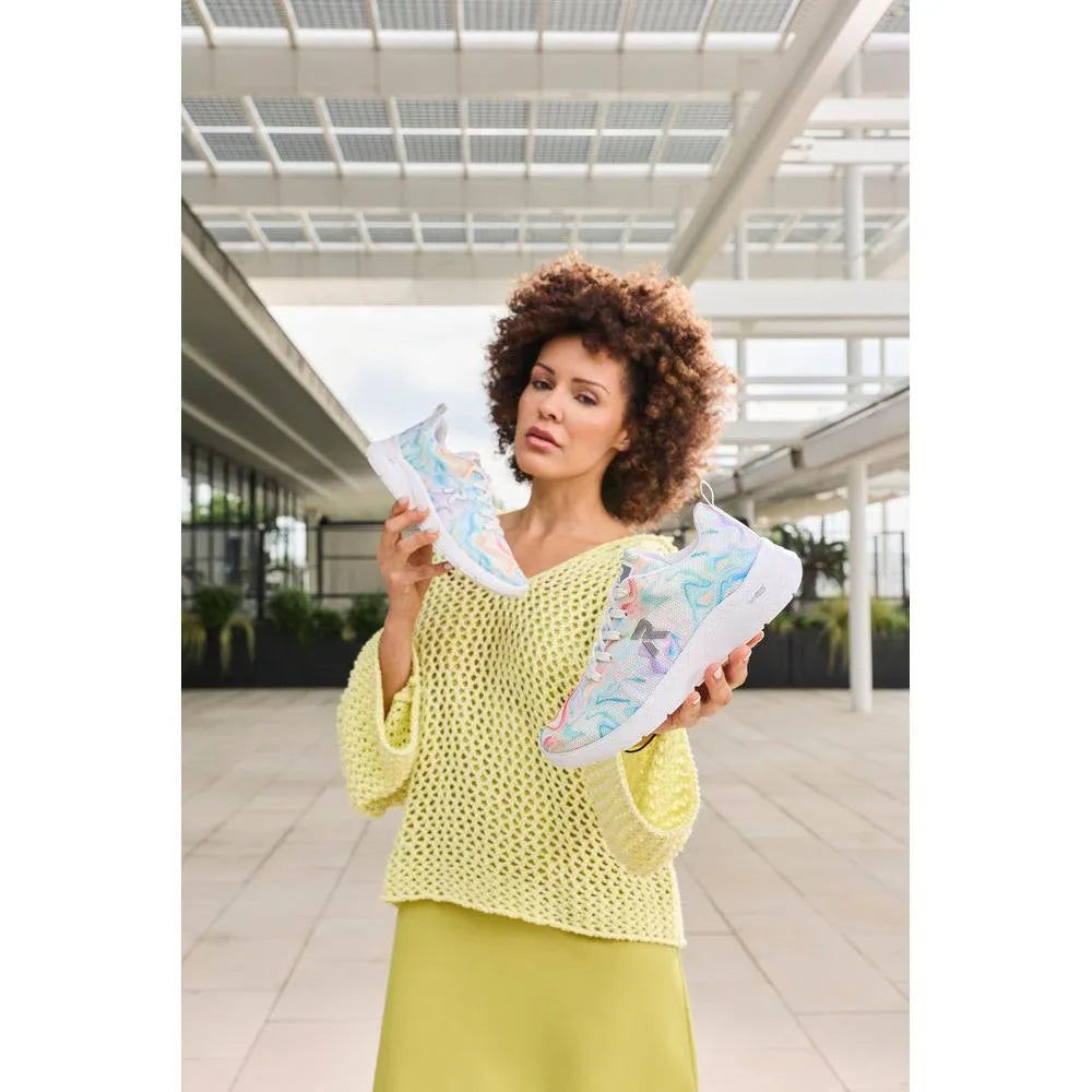 Woman holding colorful mesh vegan lace-up trainers, in a modern outdoor setting with architectural white structures.