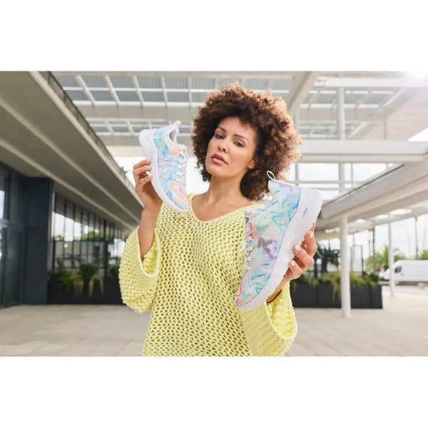 Woman holding colorful mesh vegan lace-up trainers, casual style, pastel swirls, white soles, in modern architectural setting.