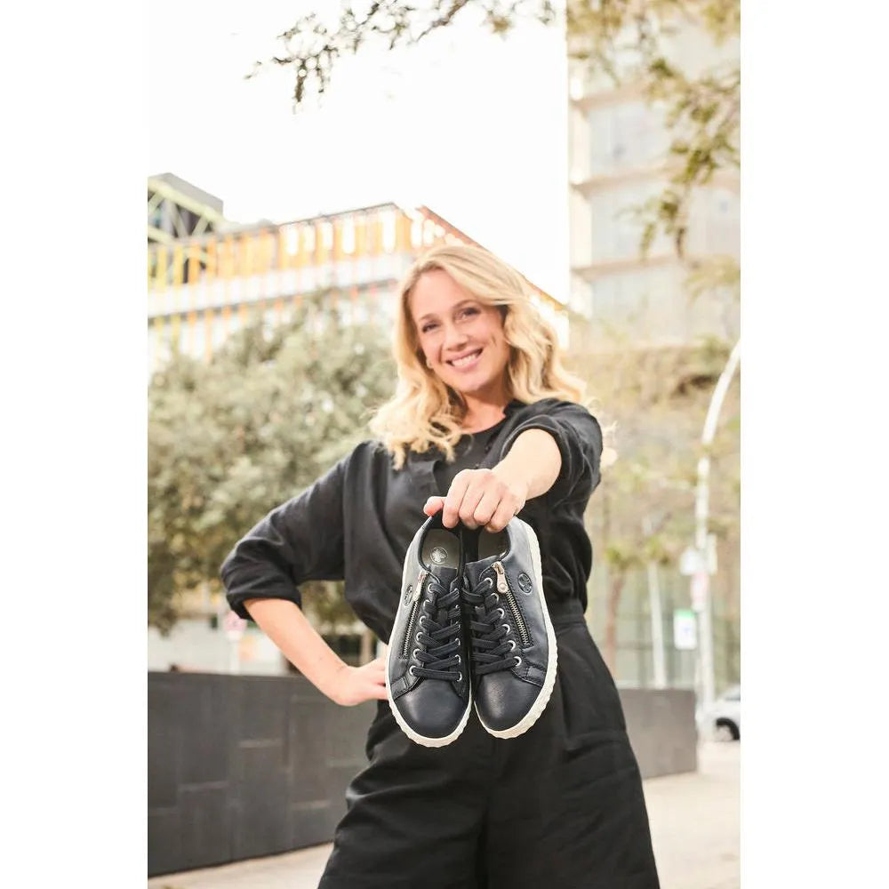 Woman holding Rieker N0900-14 Ladies Pacific Leather Zip & Lace Shoes outdoors, showcasing stylish black sneakers with zippers and laces.