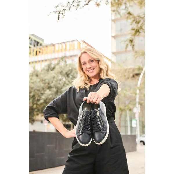 Woman holding Rieker N0900-14 Ladies Pacific Leather Zip & Lace Shoes outdoors, showcasing stylish black sneakers with zippers and laces.