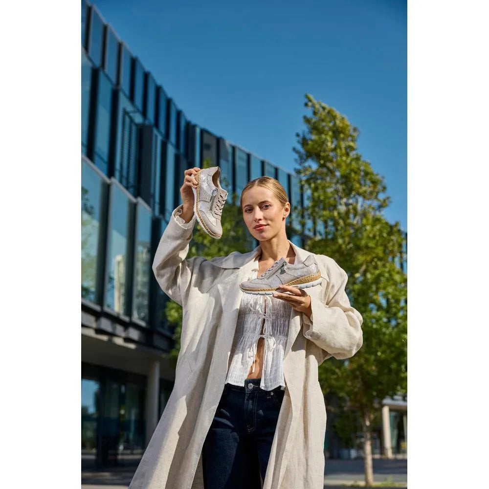 Woman holding grey Rieker N4221-90 metallic zip and lace shoes outdoors with modern building and trees in background.