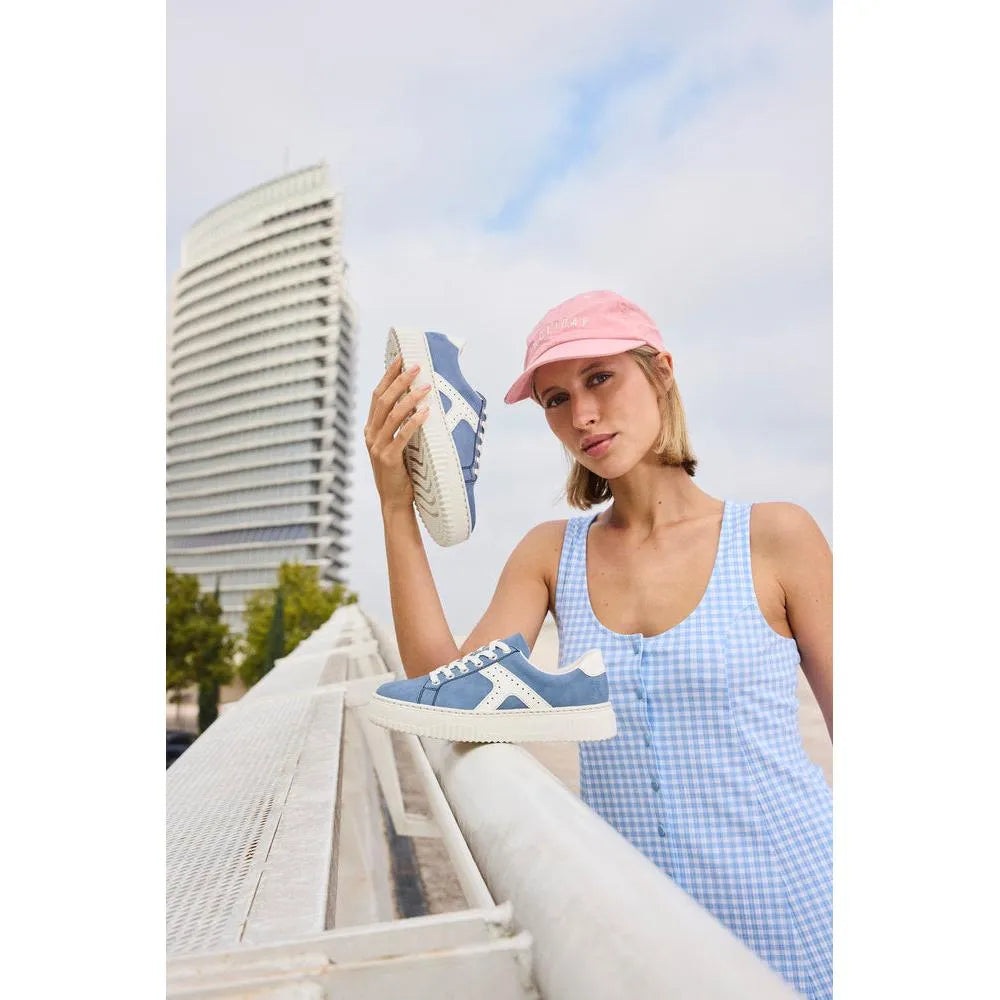 Light blue and off-white lace-up trainers with side zip detail, worn by a woman with a pink cap, outdoors against a cityscape backdrop.