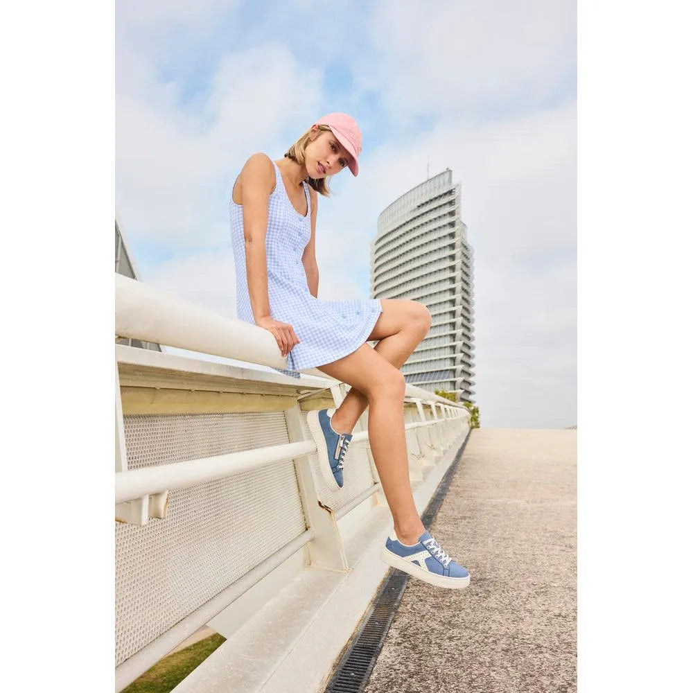 Woman sitting on a bridge in light blue and white trainers, denim dress, and pink cap, with a tall modern building in background.