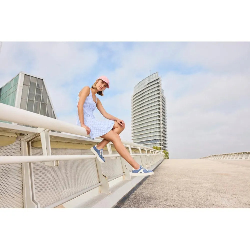 Woman in a blue and white dress, pink cap, sitting on a bridge railing in blue and white trainers, urban background with modern buildings.