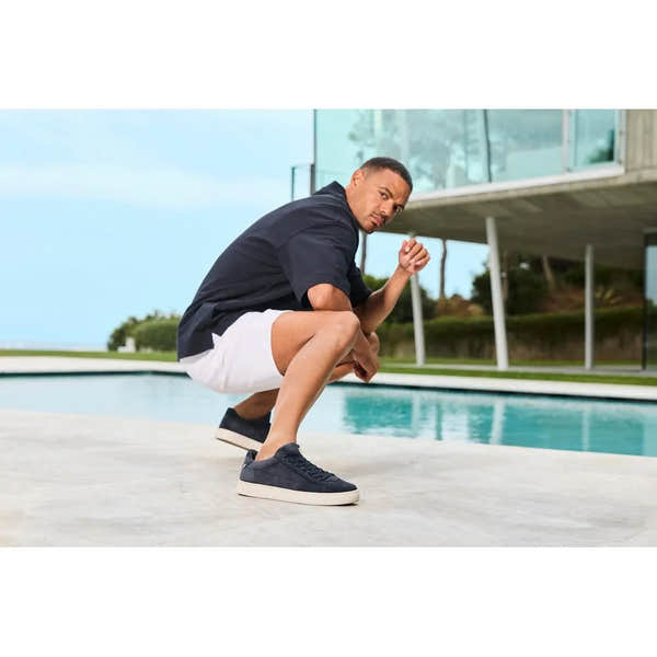 Man crouching by pool wearing navy blue suede lace-up shoes, white shorts, black top, modern building in background.