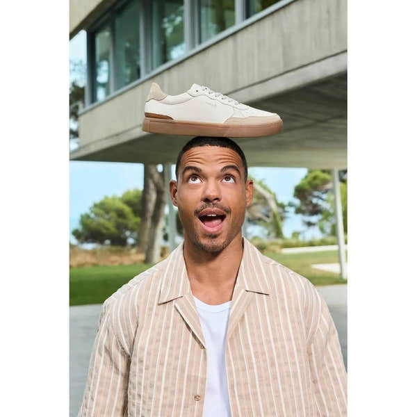 Man balancing a white leather lace-up shoe with beige accents on his head outdoors under a modern building.