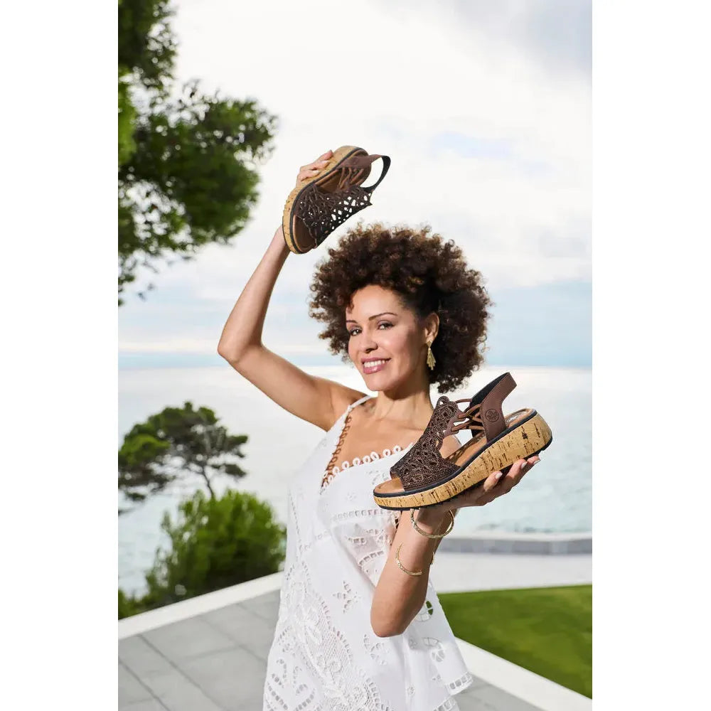 Rieker V1765-24 women's wide fit brown elasticated sandals, open-toe design, cork sole, outdoor seaside background.