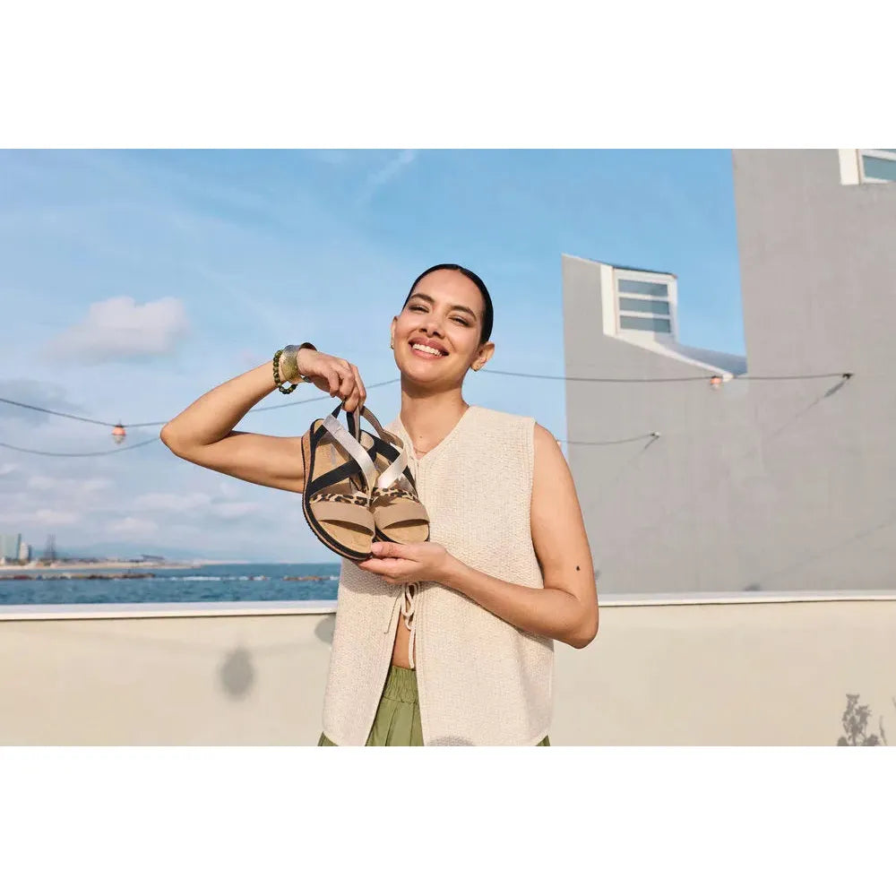 Woman smiling outdoors holding Rieker V3663-92 multi-touch fastening sandals with beige and leopard print design, casual summer footwear.