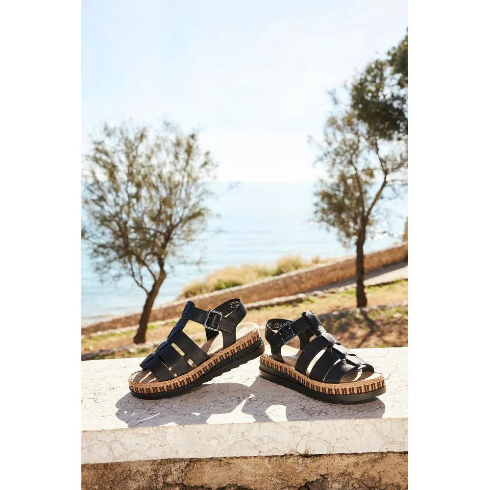 Rieker V7904-00 women's black leather touch fastening sandals outdoors by the water. Flat sandals with striped wedge soles and adjustable straps.