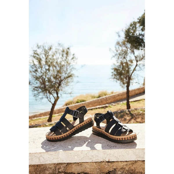 Rieker V7904-00 women's black leather touch fastening sandals outdoors by the water. Flat sandals with striped wedge soles and adjustable straps.