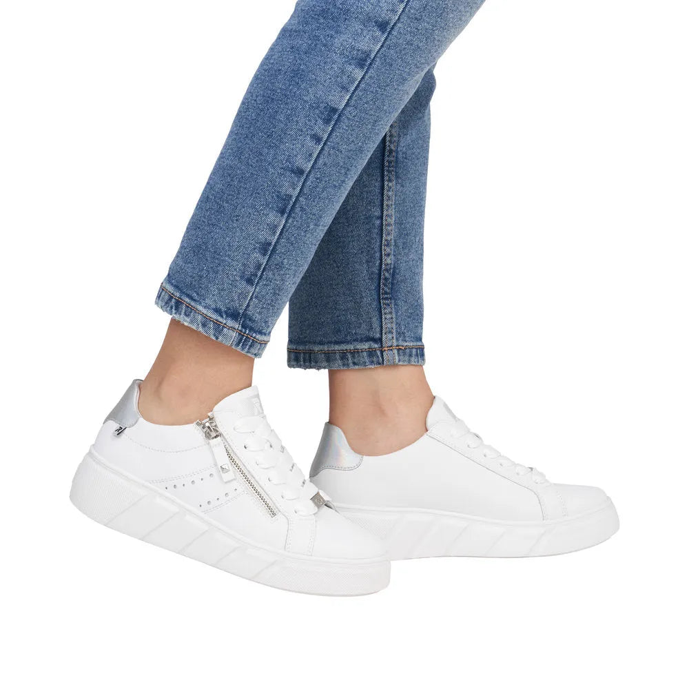 Rieker W2605-80 women's white leather sneakers with zip and laces, chunky sole, and iridescent accents, paired with blue jeans.