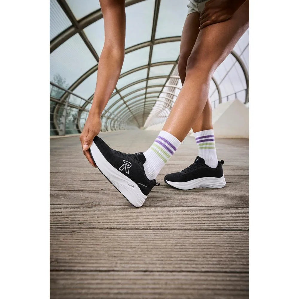 Black textile lace-up trainers with white midsole, worn with white socks featuring purple, green, and blue stripes.