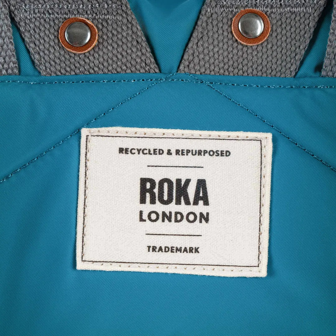 Roka London recycled nylon bag with gray shoulder straps, metal snap buttons, and white label highlighting eco-friendly material.