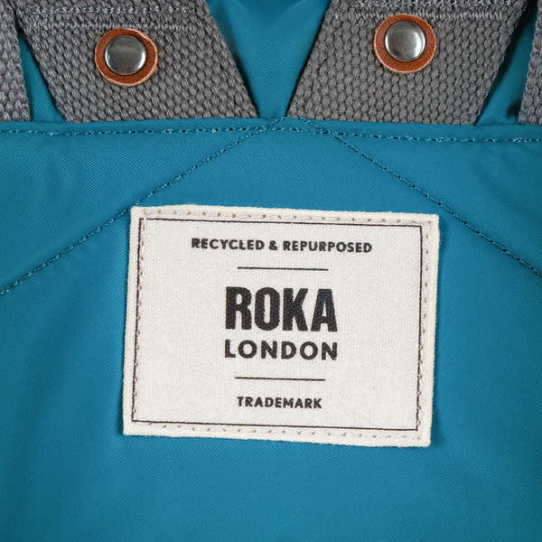 Roka London recycled nylon bag with gray shoulder straps, metal snap buttons, and white label highlighting eco-friendly material.