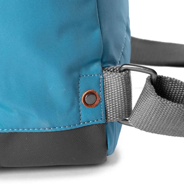 Roka Bantry B Petrol recycled nylon backpack strap detail with grey strap, metal ring, and orange-accented rivet on blue fabric.