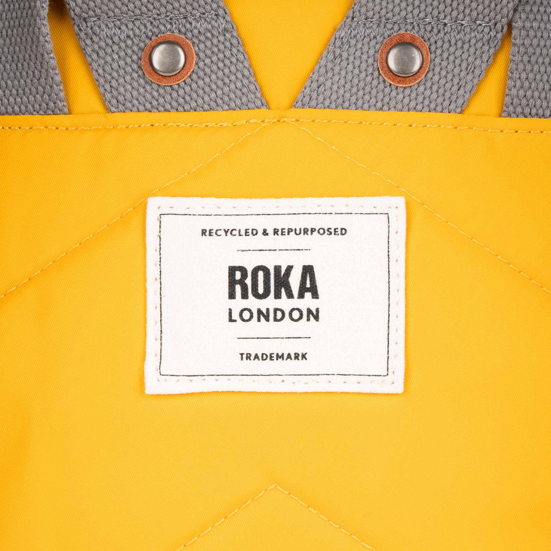 Close-up of a yellow recycled nylon backpack with gray straps, metal grommets, and a white ROKA London label.