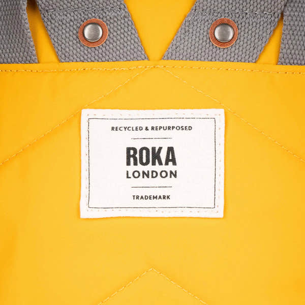 Close-up of a yellow recycled nylon backpack with gray straps, metal grommets, and a white ROKA London label.