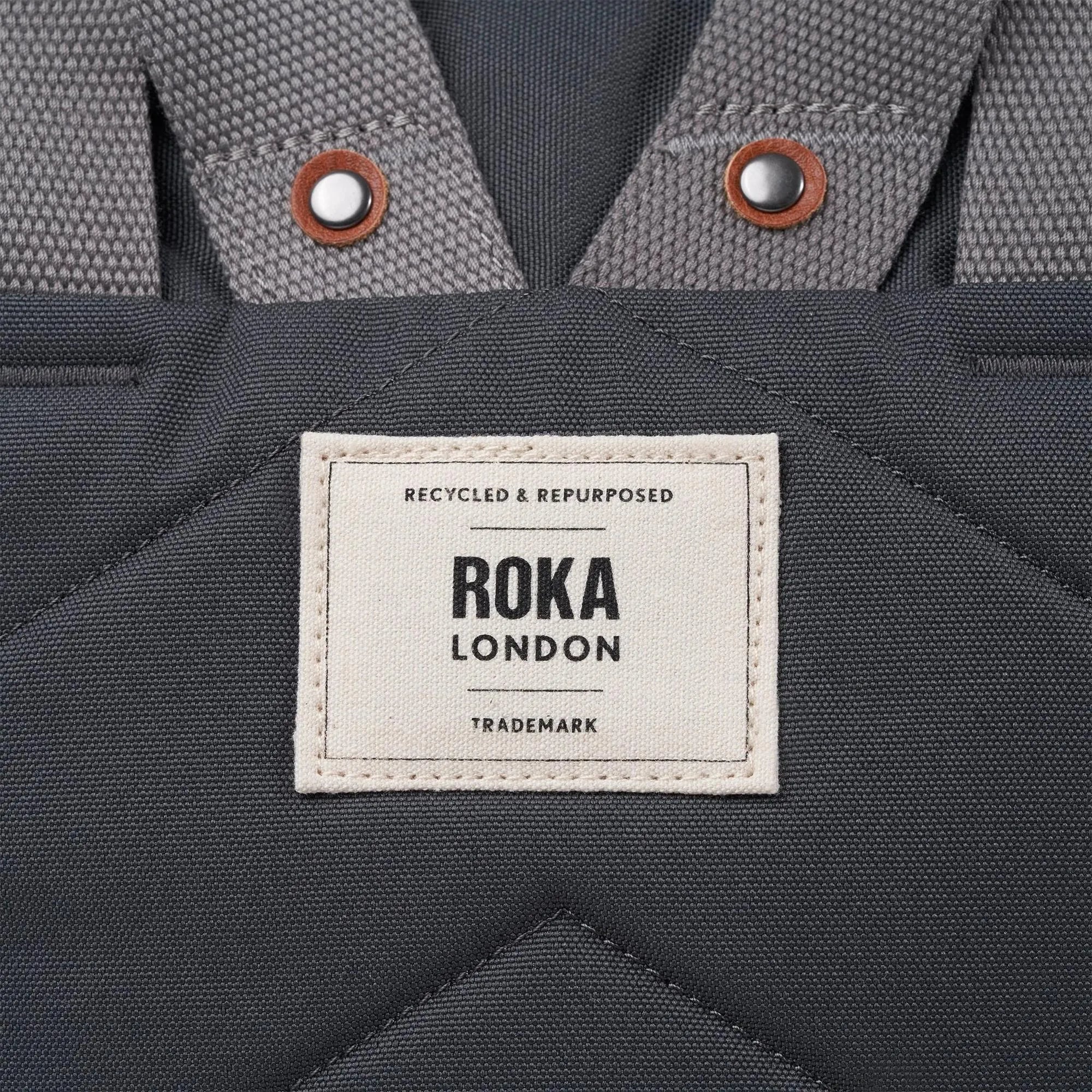 Roka Finchley A Faded Black Recycled Canvas backpack with grey straps and metal snap buttons.