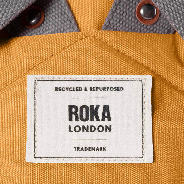 Roka London recycled canvas bag with a white label, mustard yellow fabric, and metal snap buttons.