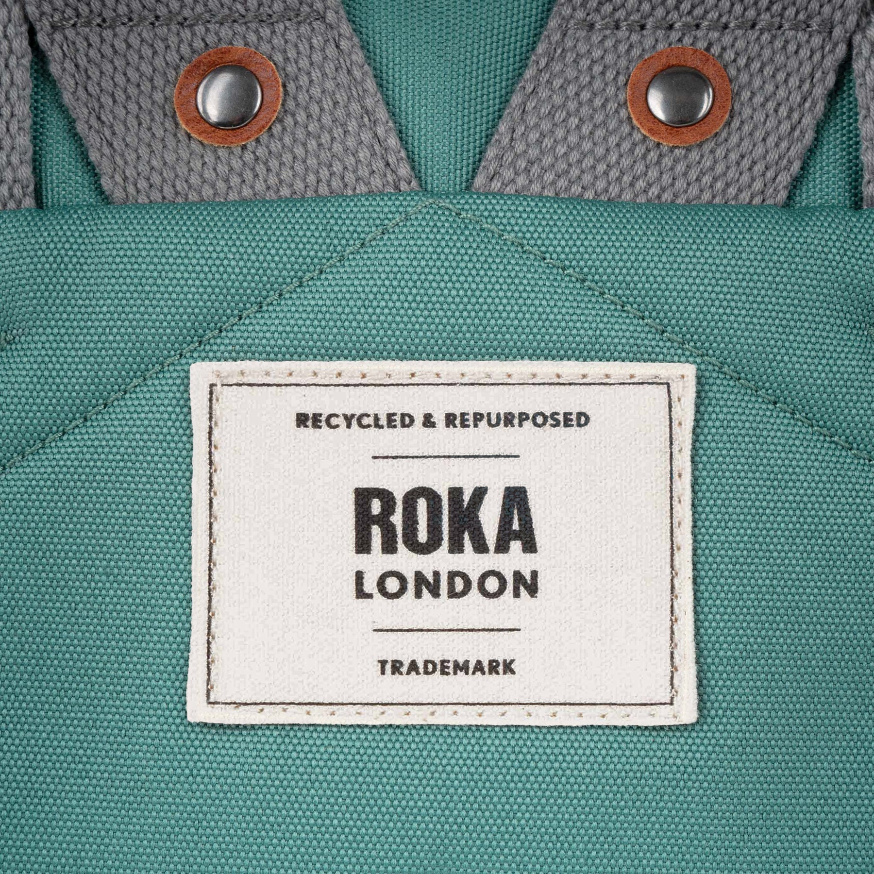 Roka Finchley A Sage Recycled Canvas backpack with brown leather and metal snap closures, close-up of label and fabric details.