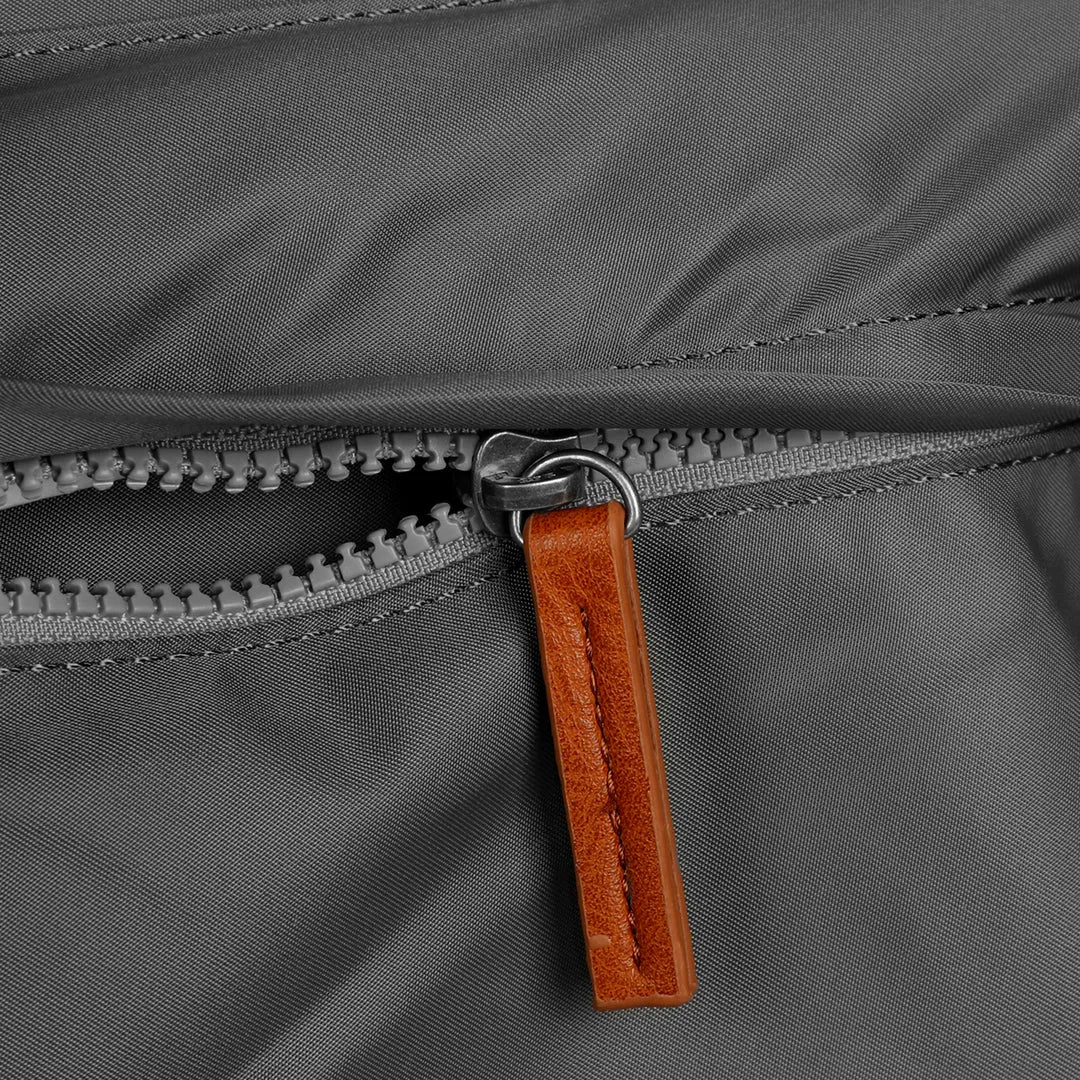 Roka Kennington B Crossbody bag in recycled nylon with graphite color, featuring a close-up of zipper and leather pull tab.