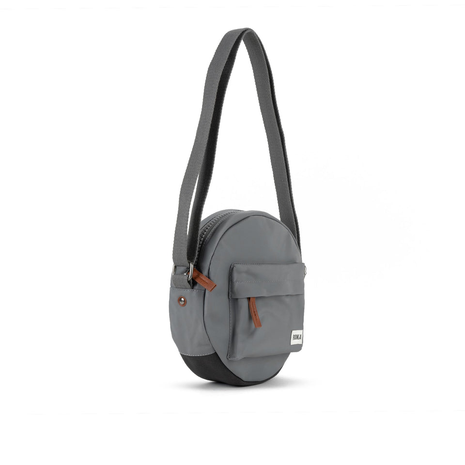 Roka Paddington B Stormy Small recycled nylon crossbody bag with front pocket, adjustable strap, and brown zipper detail.