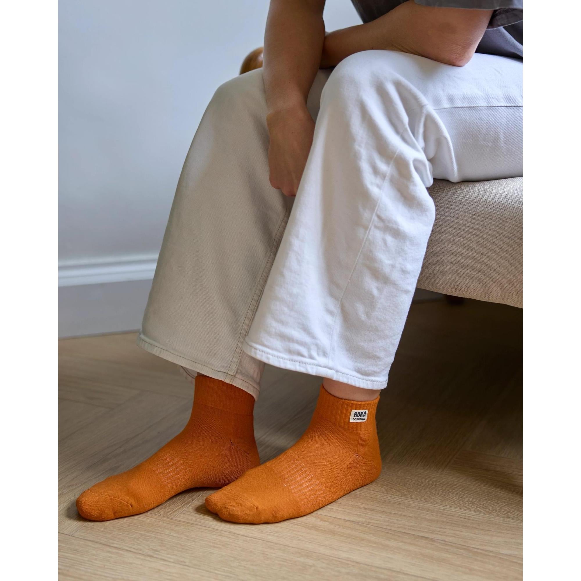 Roka Wembley burnt orange organic cotton socks, one size, worn with white pants and sitting on light wood floor.
