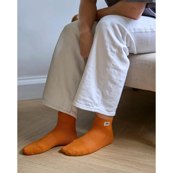 Roka Wembley burnt orange organic cotton socks, one size, worn with white pants and sitting on light wood floor.