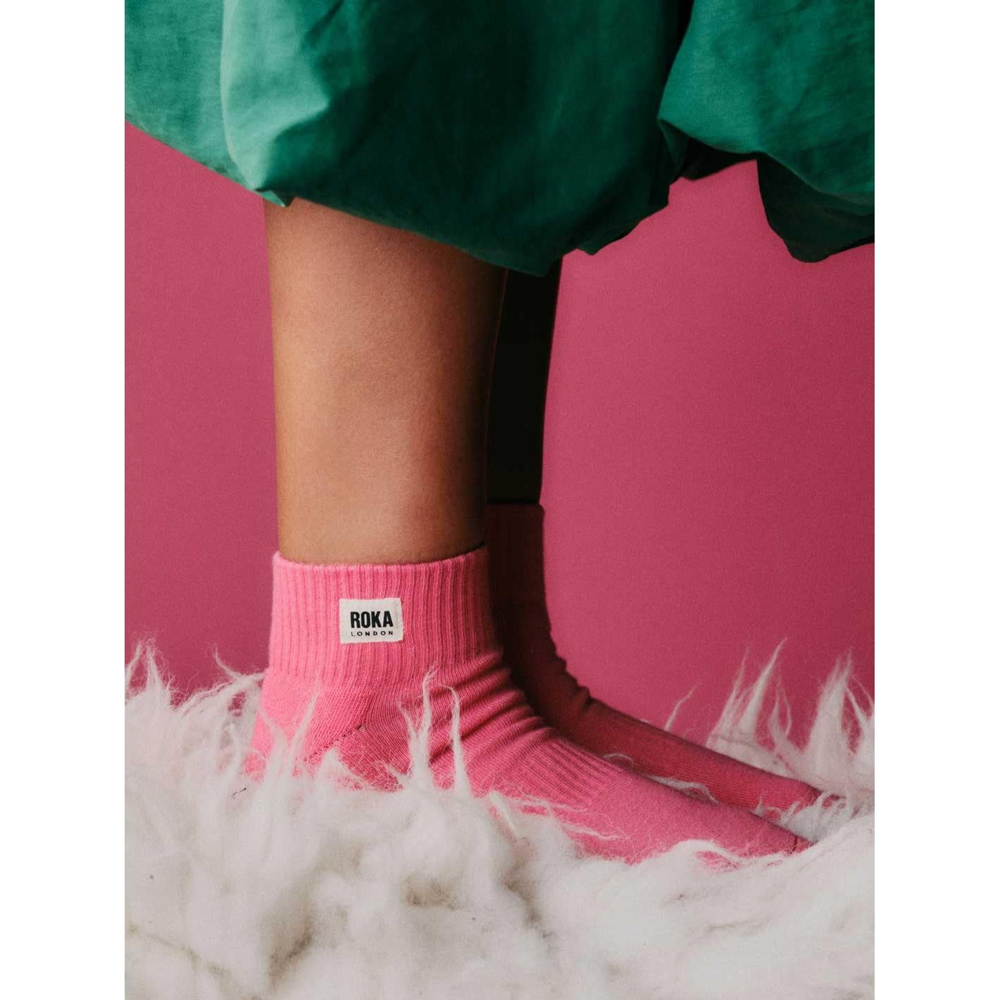 Pink cotton socks with Roka London logo, styled with a green skirt and white furry surface, ideal for casual wear.