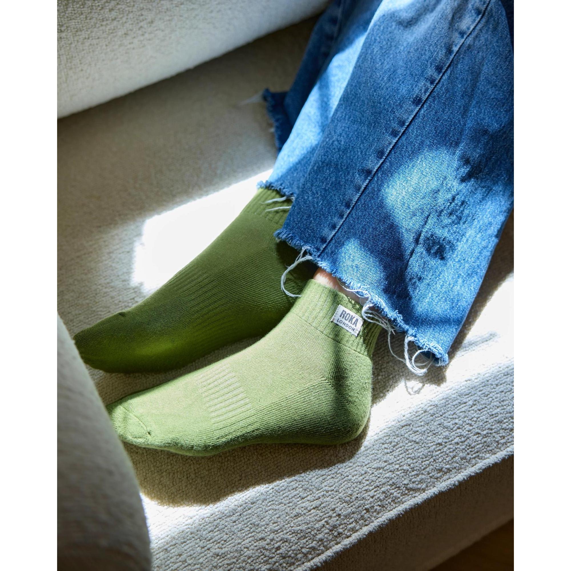 Roka Wembley ladies organic cotton socks in avocado green, worn with frayed denim jeans on a beige surface.