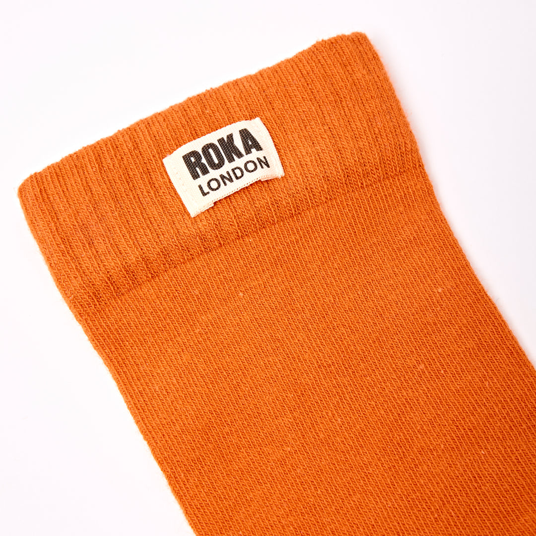 Roka London women's cotton crew sock in burnt orange, one size, with brand label, close-up view.