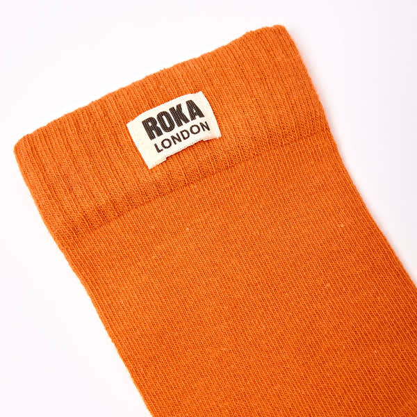 Roka London women's cotton crew sock in burnt orange, one size, with brand label, close-up view.