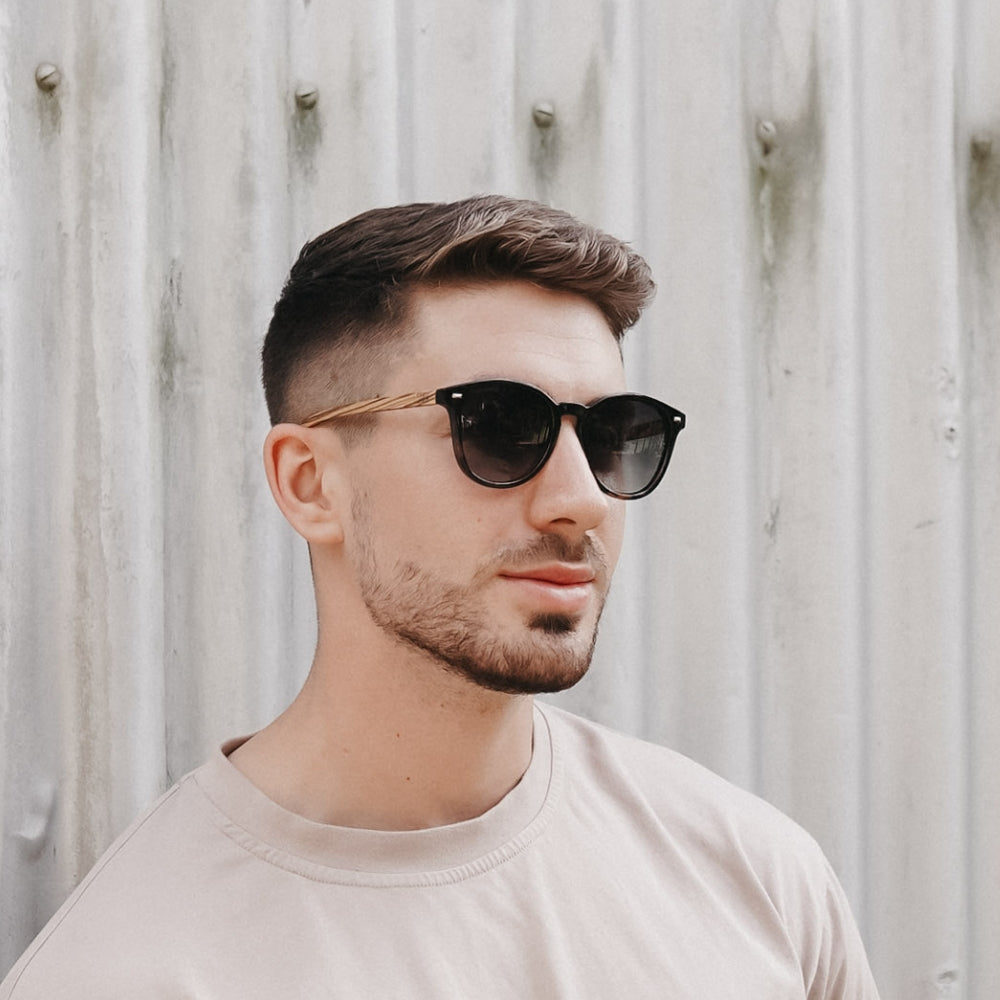 Soek Taine black tortoise sunglasses with khaki lenses and walnut arms on a man in front of a wooden wall.