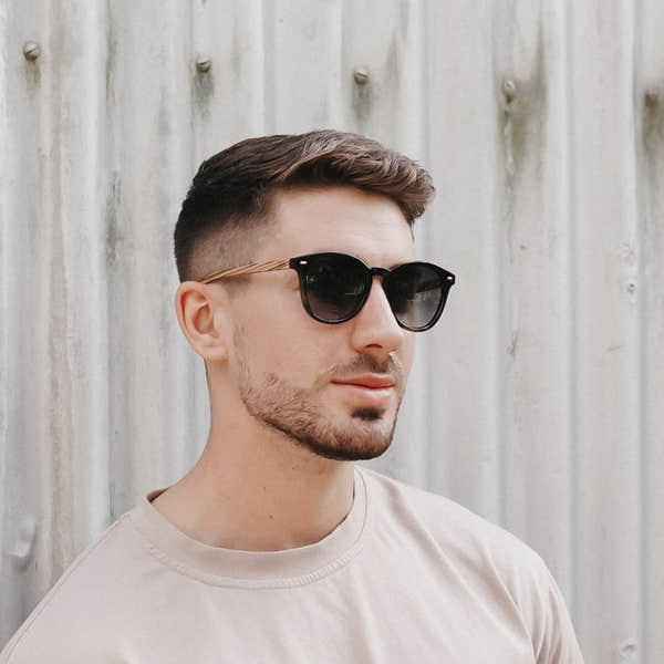 Soek Taine black tortoise sunglasses with khaki lenses and walnut arms on a man in front of a wooden wall.