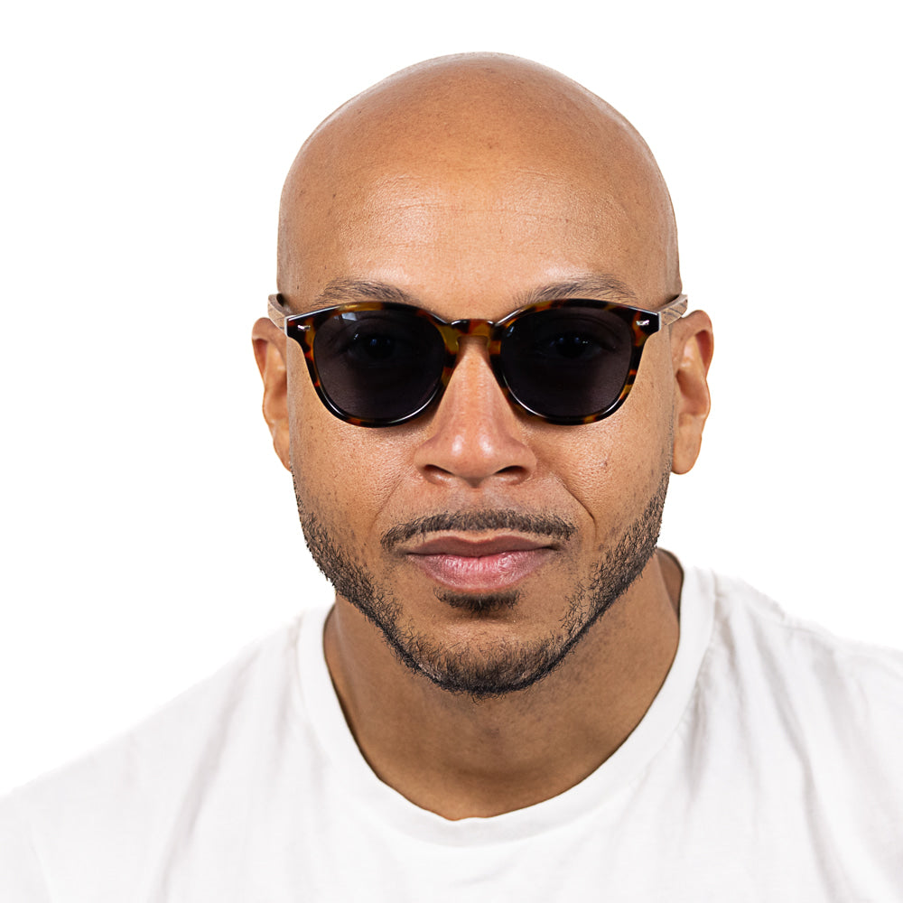 Man wearing SOEK Taine Tortoise Shell sunglasses with blue lenses and walnut arms, close-up portrait, white background.