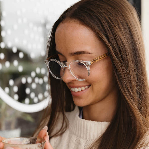 Woman wearing SOEK Wineglass Bay Blue Light Magnifying Reader glasses, smiling while holding a glass, indoors.